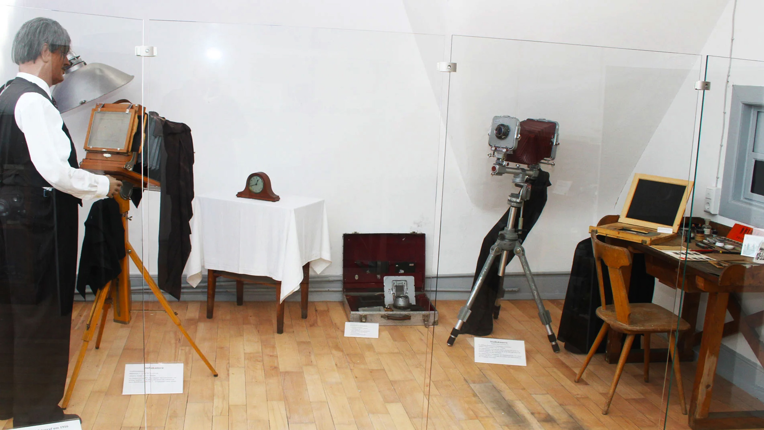 In einem Ausstellungsraum sind historische Fotografie-Geräte und ein Tisch mit fotografischen Utensilien zu sehen. Ein lebensgroßes Modell eines Fotografen steht vor einer alten Kamera, während daneben ein moderner Computer auf einem Tisch steht. Ein Wandregal zeigt eine Uhr und weitere Objekte.