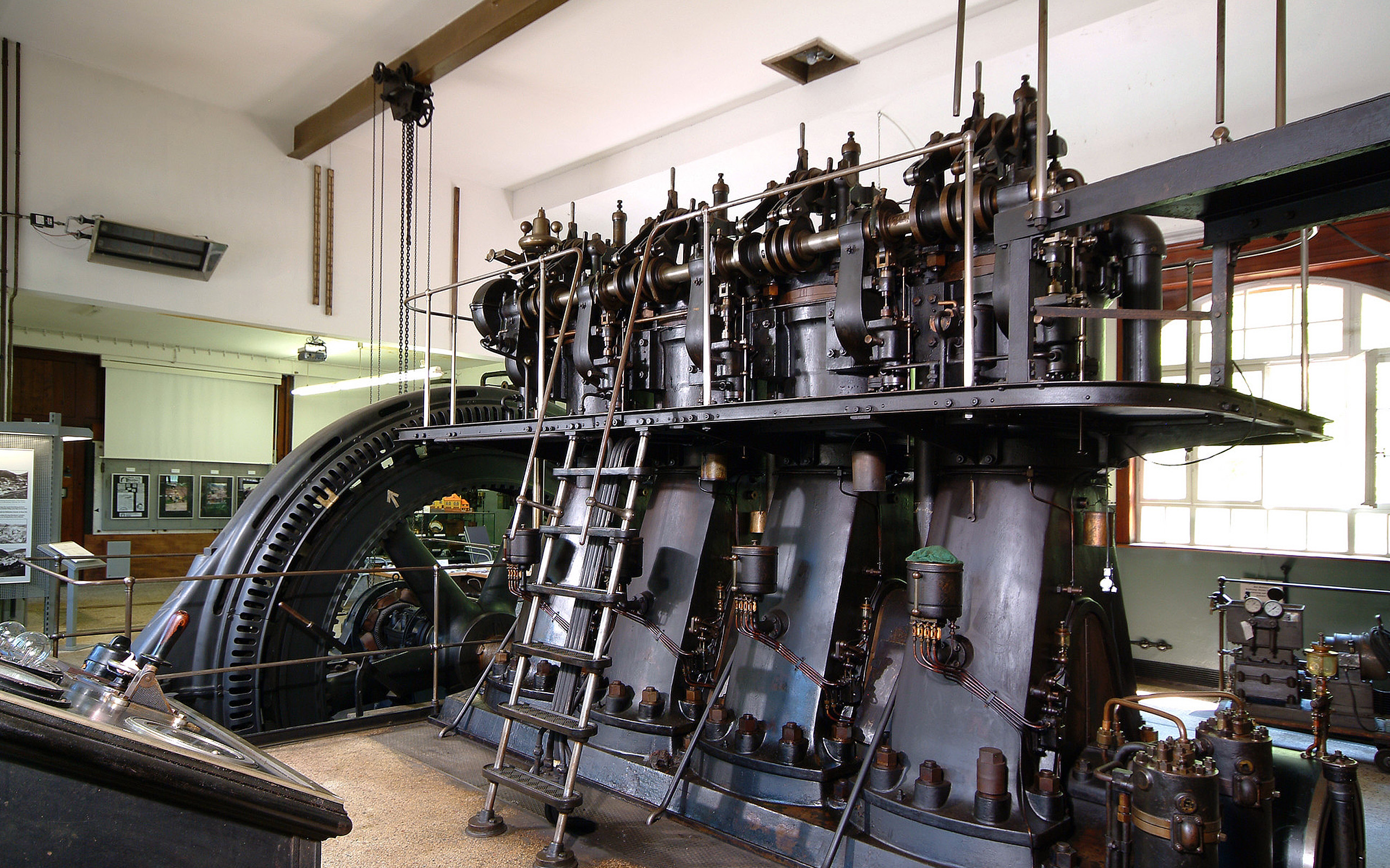 Ein großes, historisches Dampfmotor-System steht in einem Raum. Die Maschine besteht aus mehreren Zylindern, zahlreichen Rohren und einem großen Schwungrad. Eine Metallleiter führt zu einem oberen Plattformbereich. Die Szenerie vermittelt den Eindruck industrieller Technik aus vergangenen Zeiten.