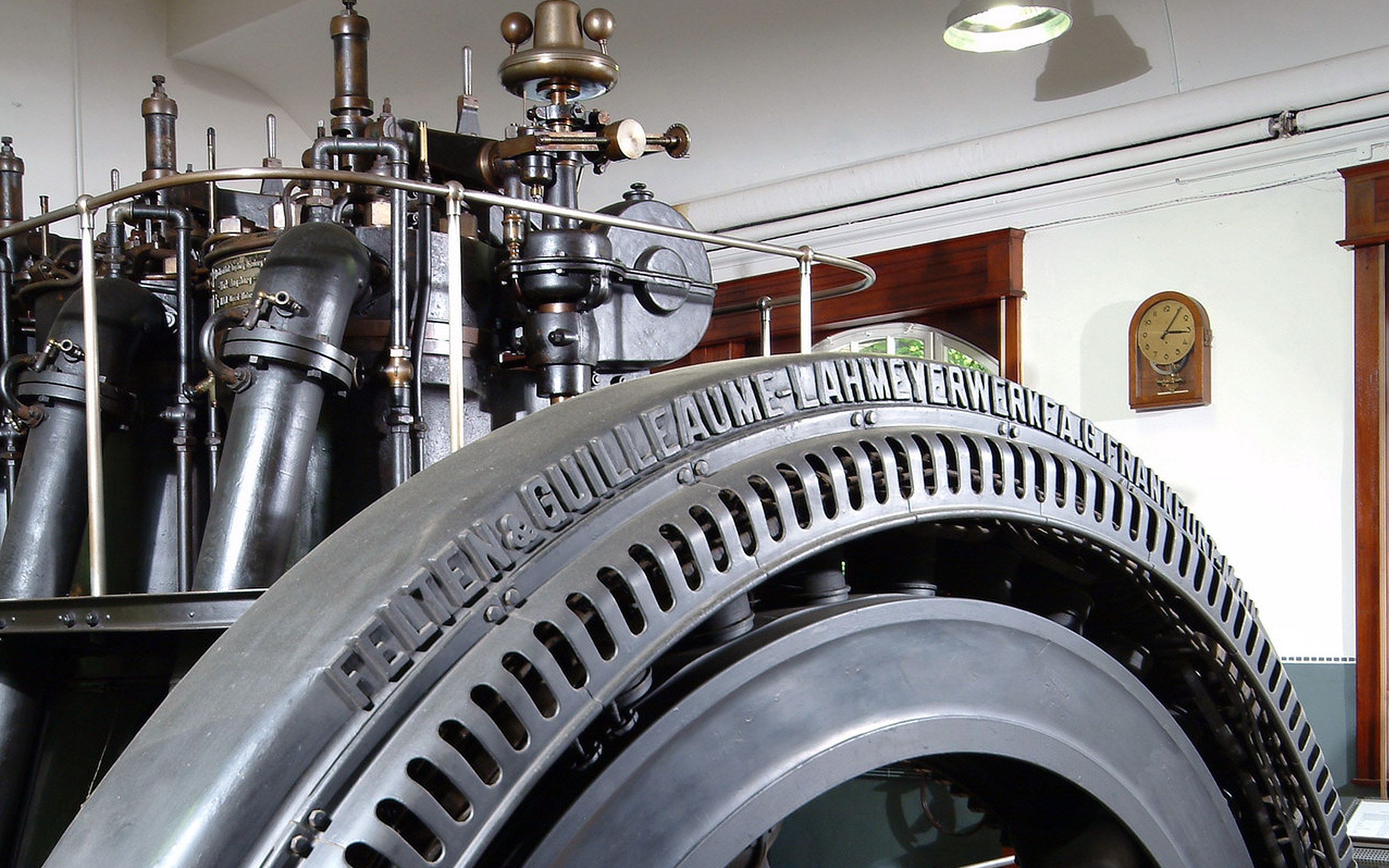Ein historischer Generator mit großen, runden, metallenen Bauteilen. Die Vorderseite zeigt einen markanten Reifen mit der Aufschrift „FELTEN & GUILLAUME - LAHMEYER WERKE A.G. FRANKFURT“ und gerippten Mustern. Im Hintergrund erkennt man Rohre und andere Maschinenbestandteile, die zur Funktionsweise des Generators beitragen.