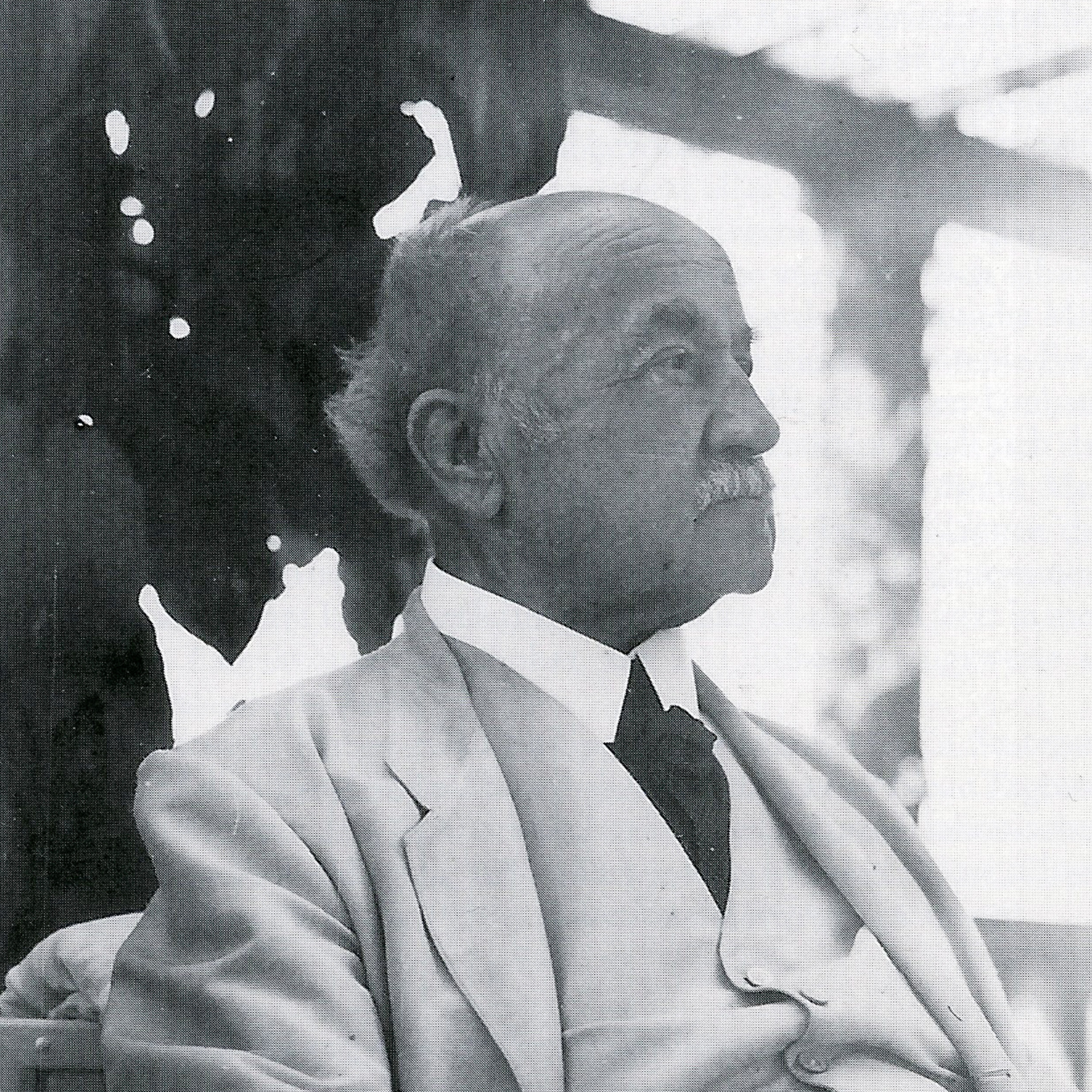 Ein älterer Mann mit einem weißen Anzug sitzt nachdenklich und schaut zur Seite. Er hat einen weißen Schnurrbart und dünnes, graues Haar. Im Hintergrund sind unscharfe, helle Formen sichtbar, die eine entspannte Umgebung andeuten.