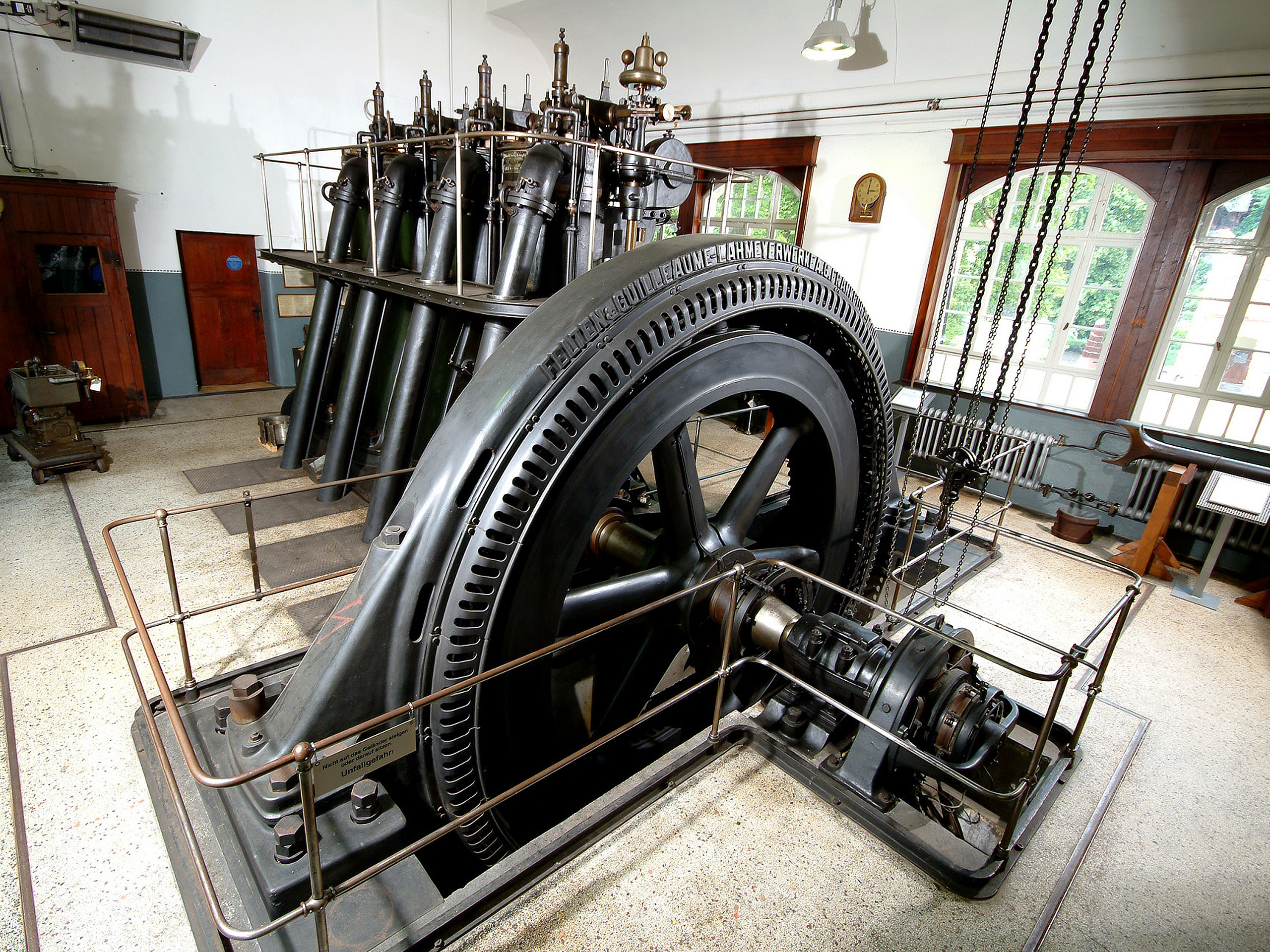 Das Bild zeigt eine große, industrielle Dampfmaschine mit einem massiven Schwungrad und zahlreichen Rohren. Der Maschinenraum ist schlicht gehalten, mit Holz- und Metallelementen. Diese historische Technik steht in einem Raum mit Fenstern, die Tageslicht hereinlassen.