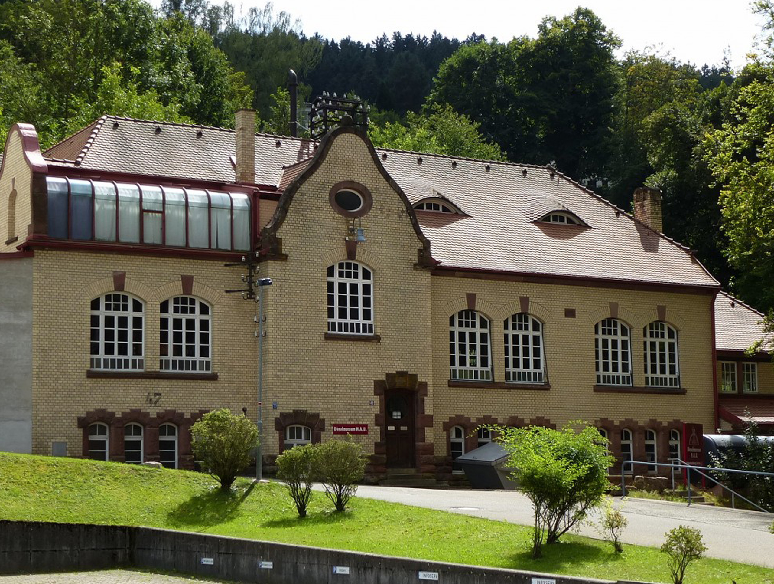 Ein historisches, zweigeschossiges Gebäude aus hellen Ziegeln mit großen Fenstern und einem roten Dach. Im Vordergrund ist eine kleine Grünfläche mit Sträuchern zu sehen. Das Gebäude ist von Bäumen umgeben und wirkt prominent in der Landschaft.