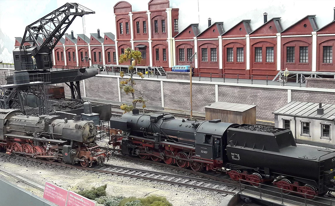 Das Bild zeigt eine Modelleisenbahn-Landschaft mit zwei Dampflokomotiven auf einem Gleis. Im Hintergrund befinden sich rote Backsteingebäude, während ein Kran über Schüttgut steht. Die Szene vermittelt eine historische Industrieatmosphäre.