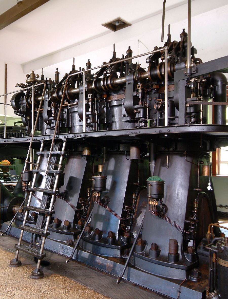 Das Bild zeigt eine große industrielle Maschine mit mehreren Zylindern und Rohren. Eine Metallleiter führt zu den oberen Teilen der Maschine. Die Struktur ist aus Stahl und wirkt robust, typischerweise für historische Dampfkraftwerke oder ähnliche Einrichtungen.