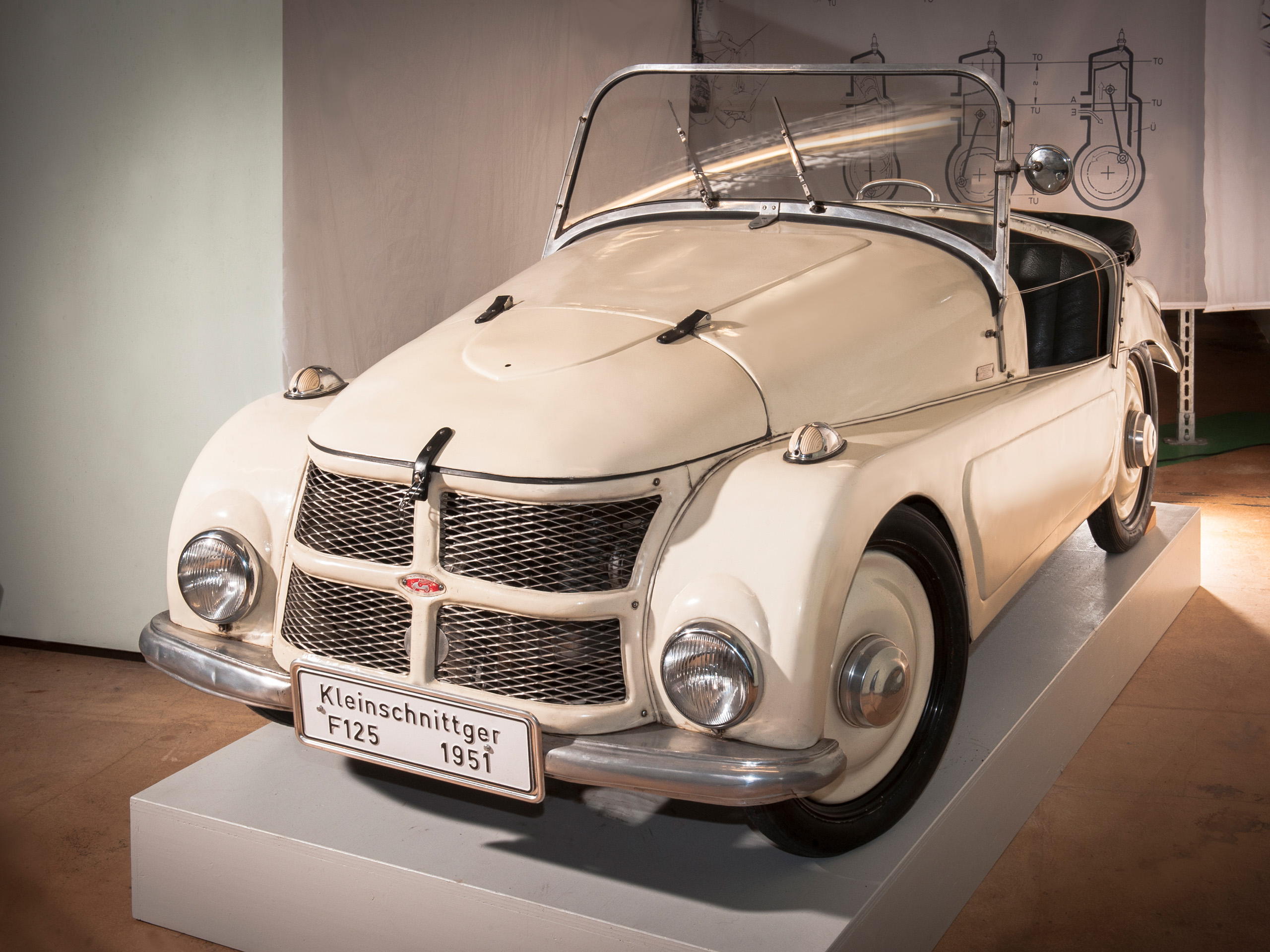 Ein historisches Auto, der Kleinschnittger F125 von 1951, steht auf einem Sockel. Das Fahrzeug ist weiß mit verchromten Details und hat ein offenes Dach. Es besitzt einen markanten Kühlergrill und runde Scheinwerfer, die seinen klassischen Charme unterstreichen.