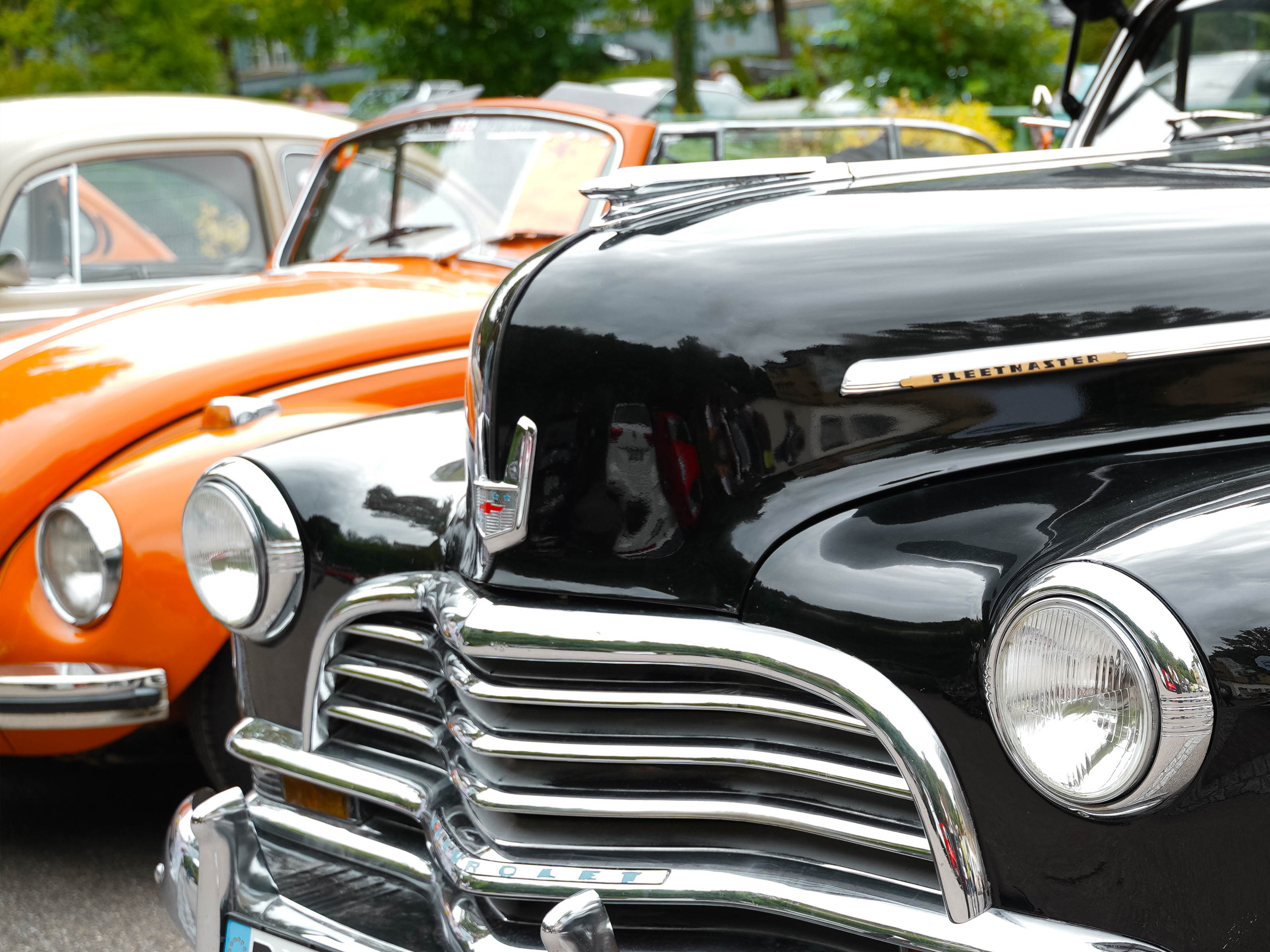 Das Bild zeigt im Vordergrund die Front eines klassischen schwarzen Autos mit glänzendem Kühlergrill und runden Scheinwerfern. Im Hintergrund sind ein orangefarbenes und ein graues Auto zu erkennen, die eine nostalgische Atmosphäre vermitteln.
