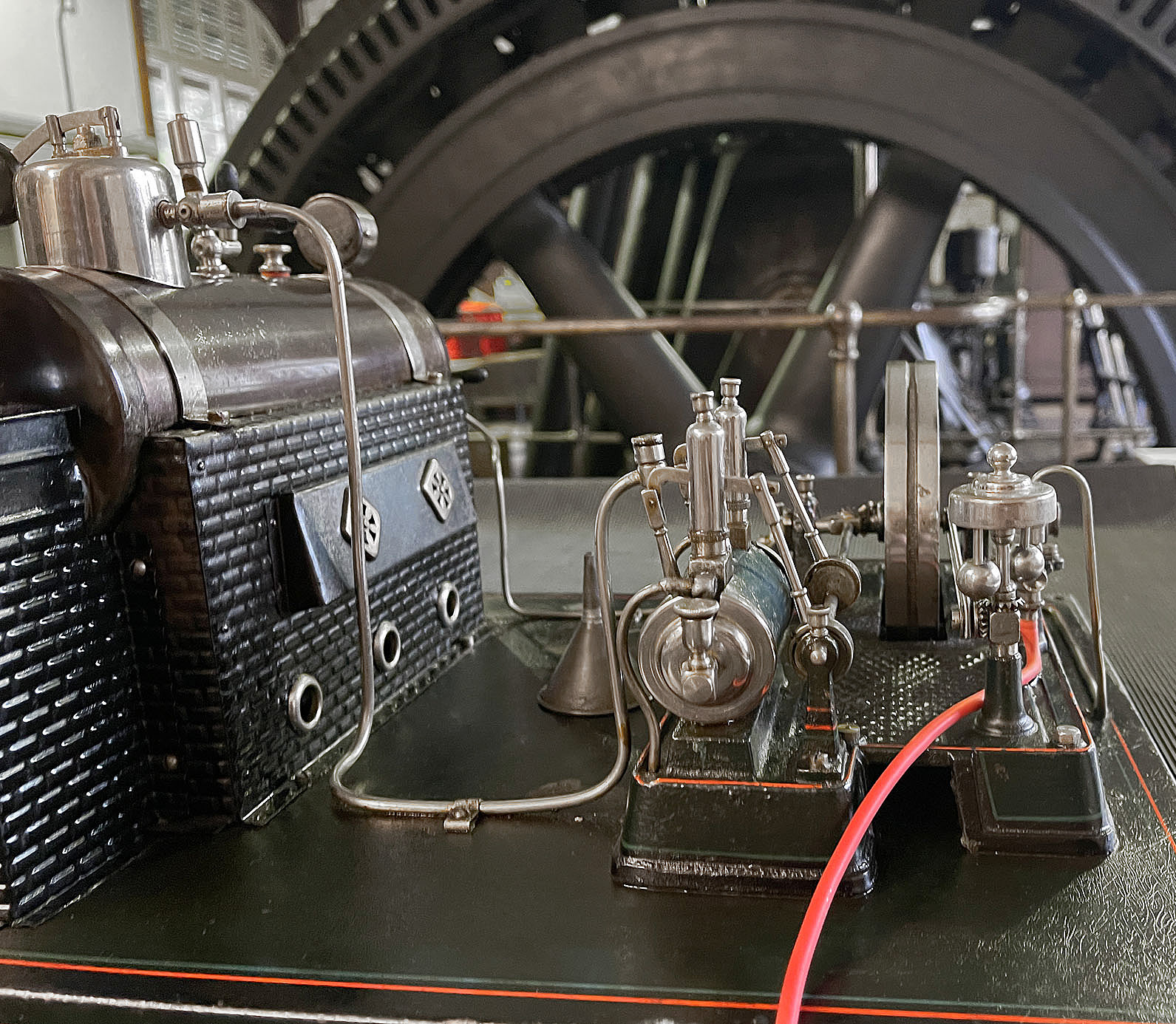 Auf dem Bild ist eine historische Modelldampfmaschine zu sehen. Sie steht vor einem großen Generator.